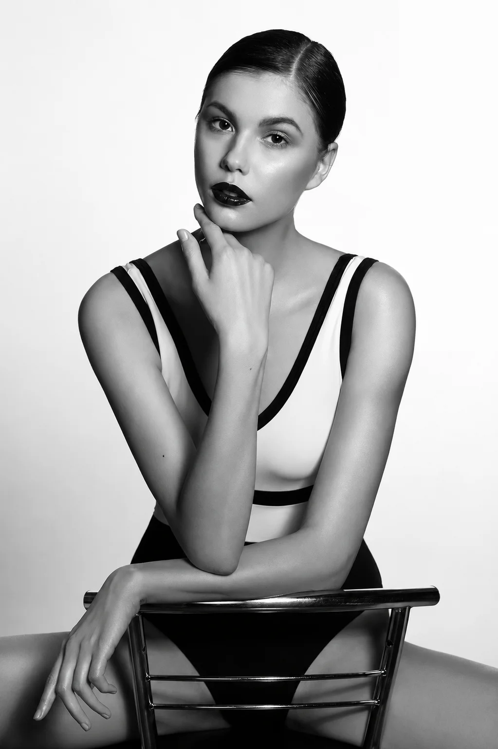 A black-and-white studio portrait of a model leaning on the back of a chair