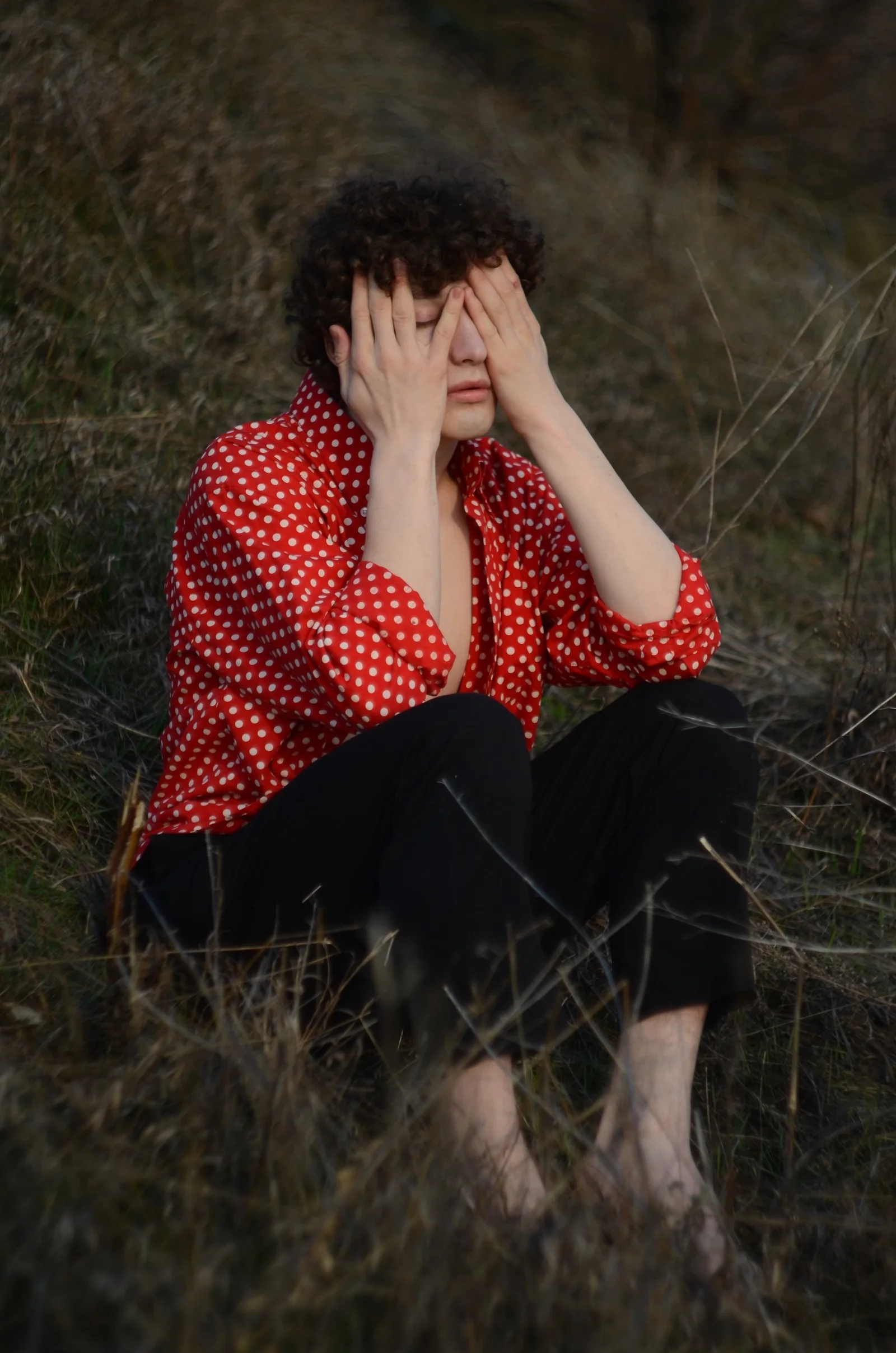 A person in a red polka-dot shirt seated in dry grass with hands over their face