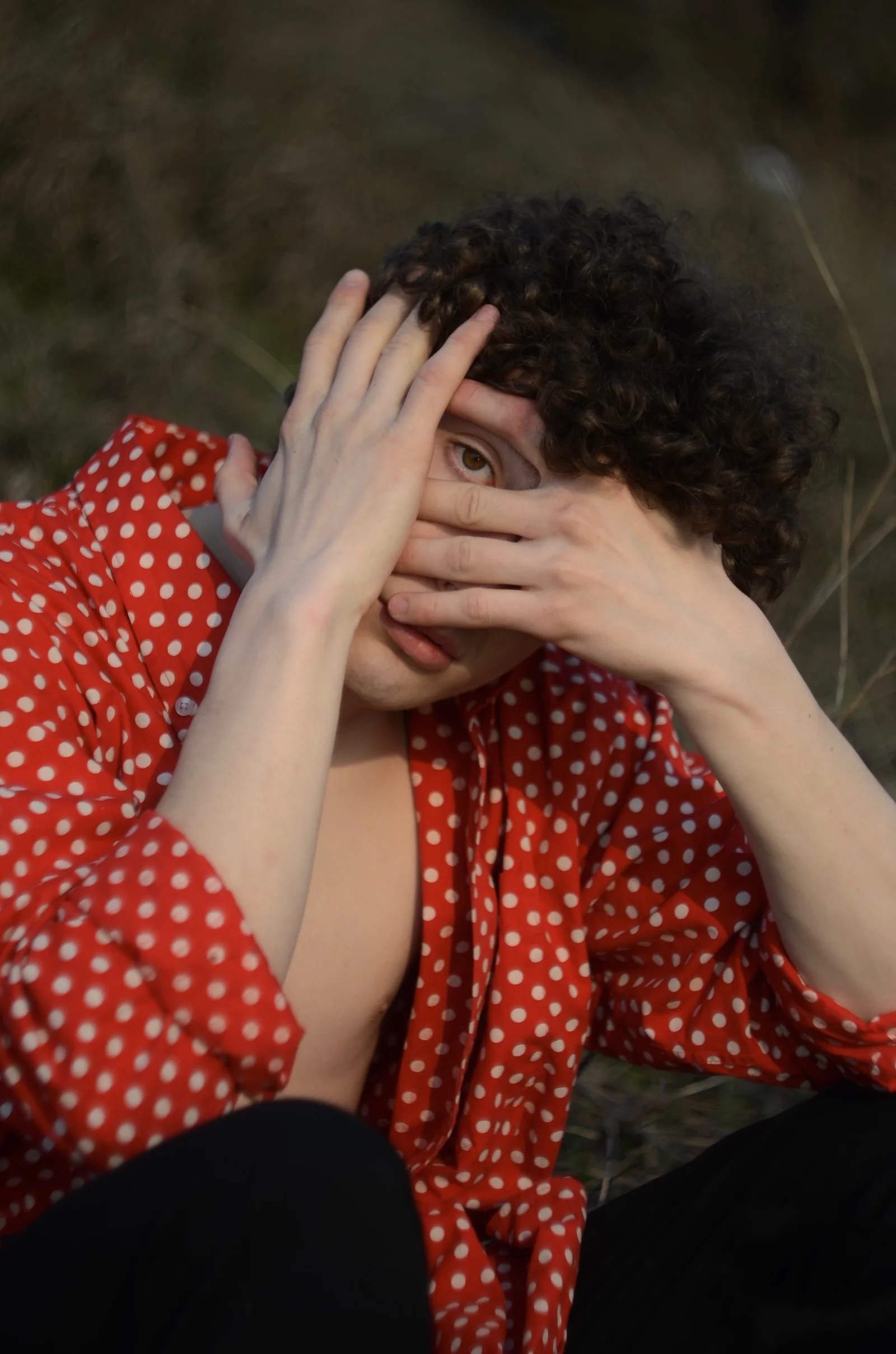 A close-up portrait in a red polka-dot shirt with an eye visible between fingers